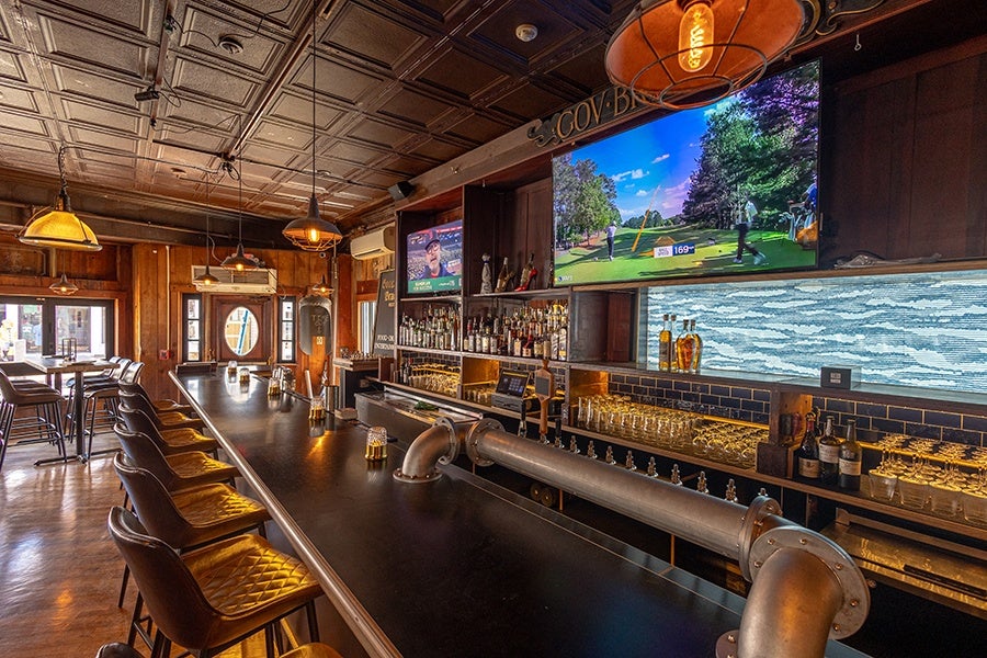 A long, dark bar counter with quilted leather bar stools lined up along one side. Behind the bar are shelves stocked with various bottles of liquor and glasses, illuminated by warm lighting. Two large flat-screen TVs are mounted above the shelves, one showing a golf game. The ceiling has a vintage tin tile design with multiple hanging pendant lights casting a cozy glow. The overall atmosphere is warm and inviting with wood-paneled walls and a polished wooden floor.