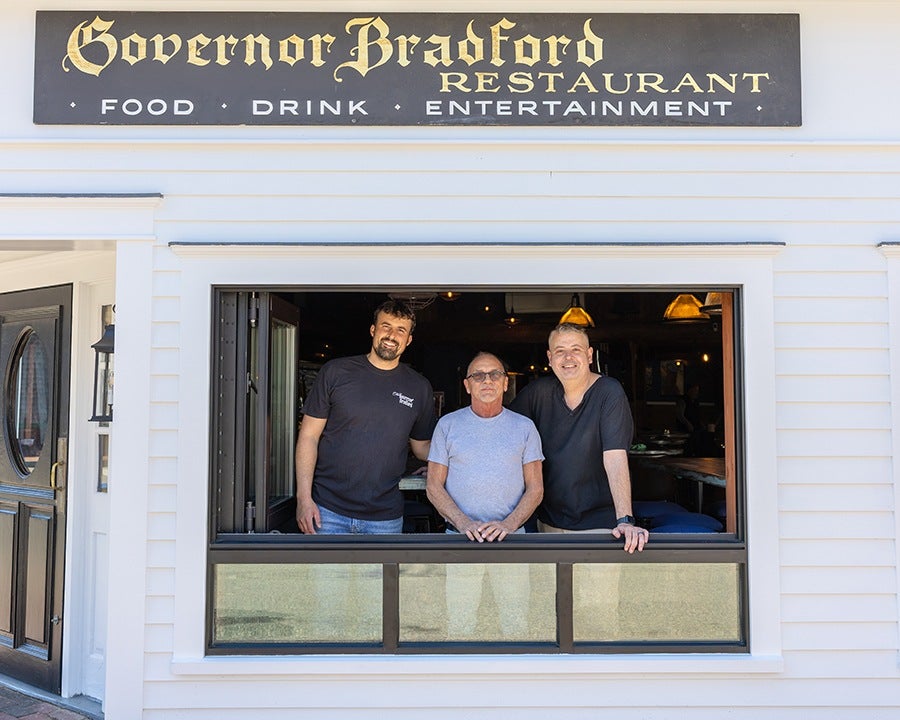 Three men stand inside a restaurant, leaning on a large open window. The restaurant exterior is white with a black door to the left. Above the window is a black sign with gold and white lettering that reads "Governor Bradford Restaurant" and "Food Drink Entertainment." The interior is dimly lit with warm hanging lights.