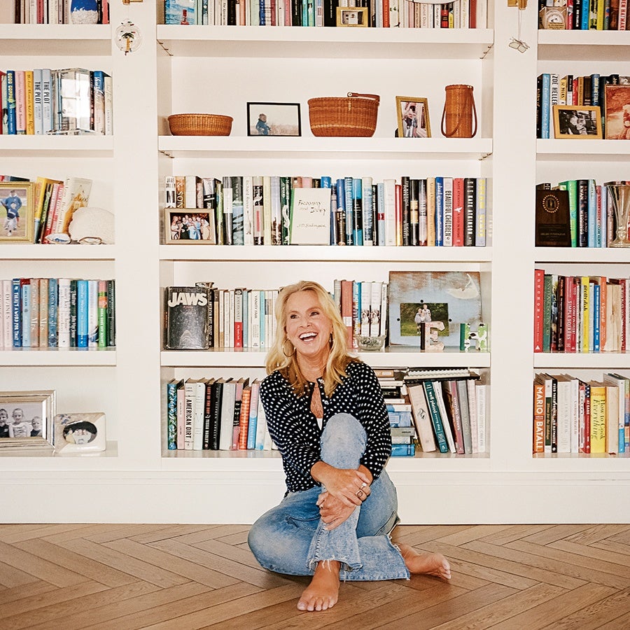 A smiling woman with blonde hair, wearing a black and white polka dot top and light blue jeans, sits barefoot on a wooden herringbone floor in front of a white bookshelf filled with books, framed photos, and decorative items.