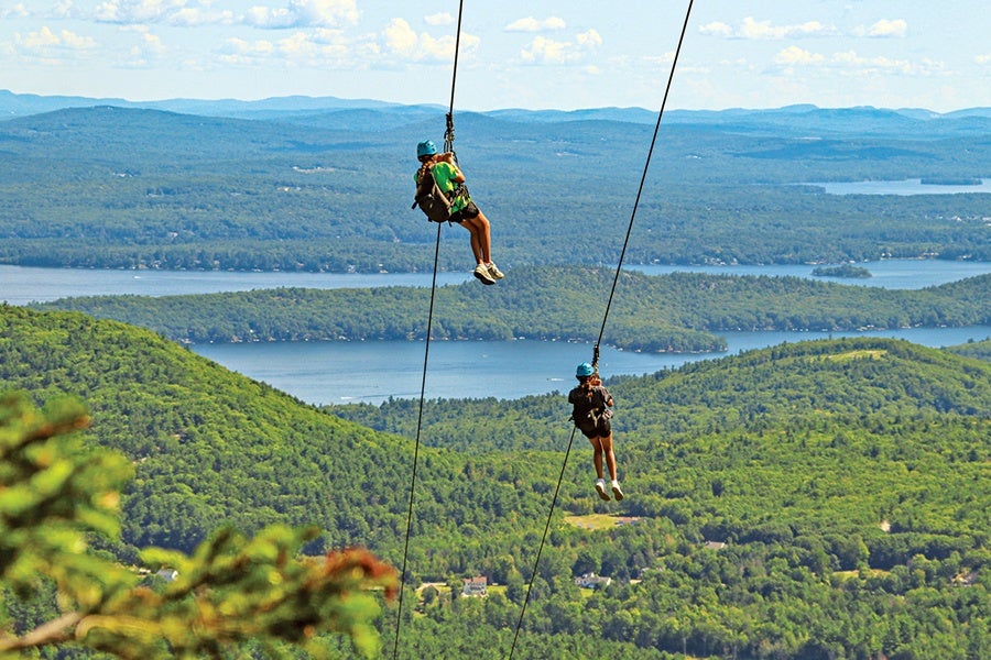 Two people wearing helmets and harnesses are ziplining over a lush green forest with a large lake and distant mountains in the background under a partly cloudy sky.