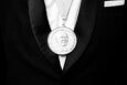 A close-up of a person wearing a black tuxedo with a white shirt and a white pocket square, displaying a large round medal on a ribbon around their neck. The medal features a relief portrait of a man and the text "JAMES BEARD FOUNDATION AWARD FOR EXCELLENCE" around the edge.