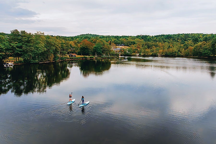 Two people paddleboarding on a calm lake surrounded by dense green and autumn-colored trees under a cloudy sky.