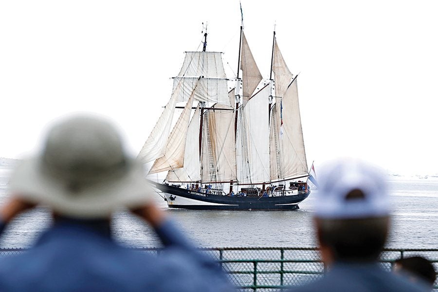 A large sailing ship with multiple white and beige sails is on the water, viewed from behind two people wearing hats who are watching it. The ship has a dark hull and is named "Oosterschelde." The scene appears to be on a calm body of water with a bright, overcast sky.