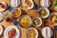 A wooden table set with various dishes including a bowl of risotto, a plate of scallops with corn and sauce, a salad with cherry tomatoes and cucumbers, a plate of roasted vegetables, a bowl of Brussels sprouts with cheese, a dish with a piece of cooked fish in broth, and a plate with three rectangular pastries topped with red garnish. There are also blue water glasses, wine glasses with red and white wine, white plates with folded napkins, and silverware arranged around the table.