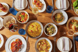 A wooden table set with various dishes including a bowl of risotto, a plate of scallops with corn and sauce, a salad with cherry tomatoes and cucumbers, a plate of roasted vegetables, a bowl of Brussels sprouts with cheese, a dish with a piece of cooked fish in broth, and a plate with three rectangular pastries topped with red garnish. There are also blue water glasses, wine glasses with red and white wine, white plates with folded napkins, and silverware arranged around the table.