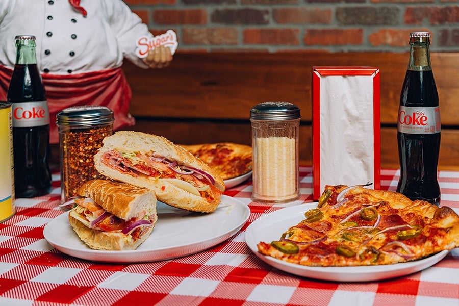 Two sandwiches with deli meats, cheese, tomato, and onions on a white plate, a slice of pizza with jalapeños and onions on another white plate, two glass bottles of Diet Coke, a container of grated cheese, a container of crushed red pepper flakes, and a red and white napkin holder on a red and white checkered tablecloth.