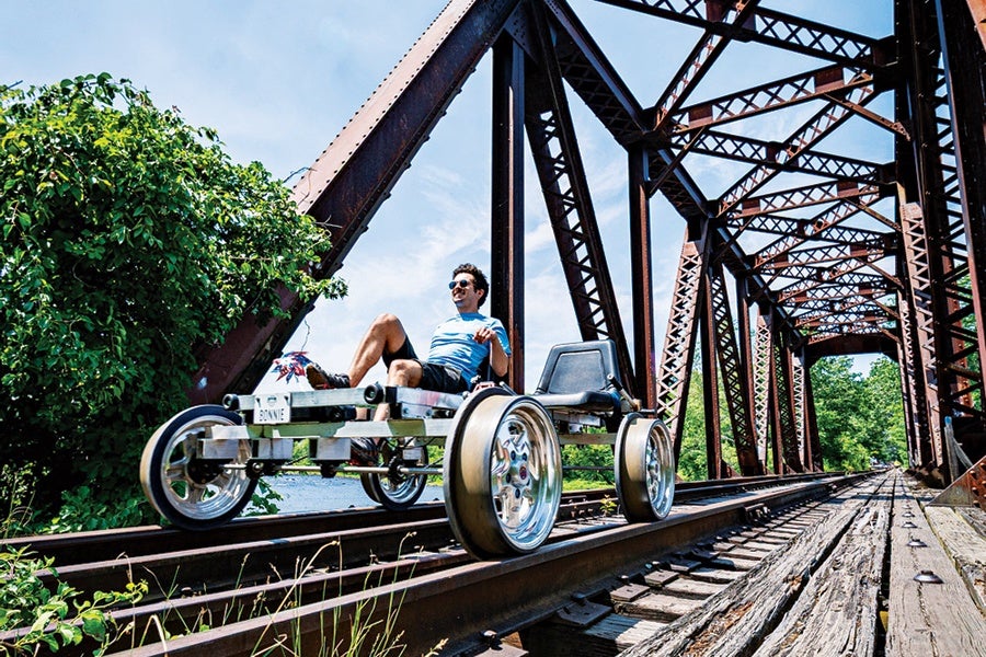 A man wearing sunglasses and a blue shirt is riding a custom-built, four-wheeled rail vehicle on railroad tracks. The vehicle has a metal frame, large shiny wheels, and a black seat. It is positioned on an old iron railway bridge with a lattice structure, surrounded by greenery and a partly cloudy sky.