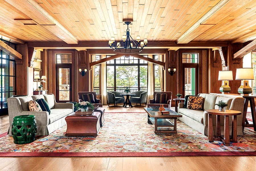 Spacious living room with wooden ceiling and walls, featuring two beige sofas with patterned cushions facing each other across a large floral area rug. Between the sofas are two coffee tables, one wooden and one with a glass top, decorated with books and small plants. The room has large windows and glass doors letting in natural light, with a seating area of four armchairs and a round table near the windows. Two table lamps with beige shades sit on a side table next to one sofa, and a green ceramic garden stool is beside the other. A black chandelier hangs from the ceiling in the center of the room.