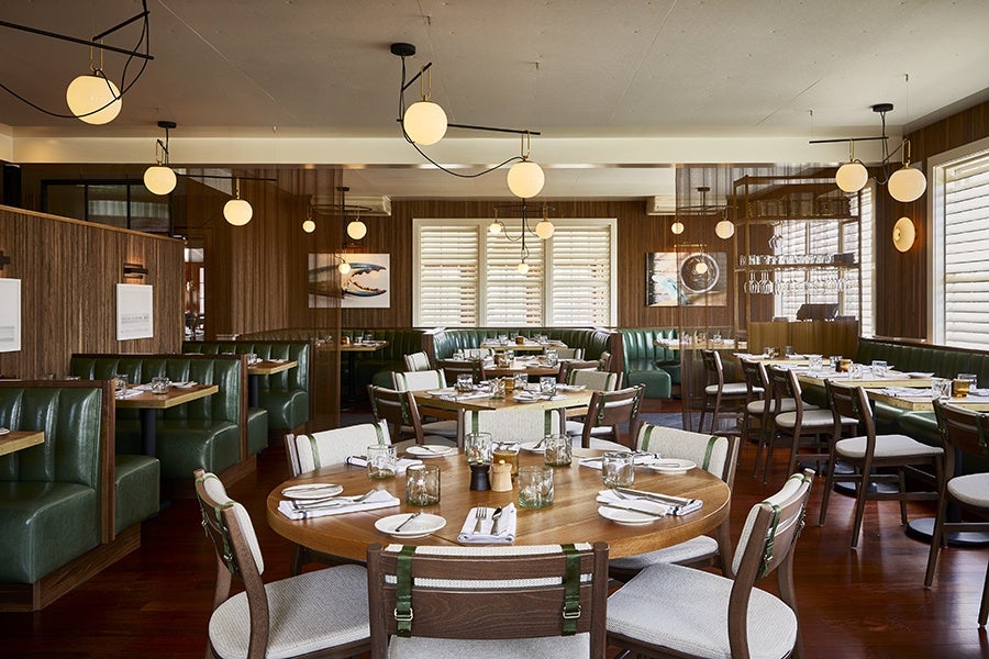 A modern restaurant interior featuring wooden floors and walls with green leather booth seating along the left and back walls. Several round and rectangular wooden tables are set with white plates, napkins, glassware, and cutlery. Light beige cushioned chairs surround the tables. Multiple spherical pendant lights hang from the ceiling, providing warm lighting. Large windows with white shutters allow natural light to enter the space. Artwork is displayed on the walls, and the overall atmosphere is clean and inviting.