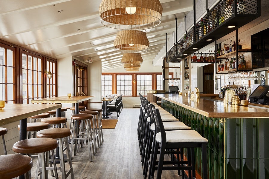 A modern bar interior with a long wooden countertop and green tiled front. High black bar stools with light cushions line the bar. On the left, there are tall wooden tables with round brown cushioned stools. Large windows with wooden frames let in natural light. Woven basket-style pendant lights hang from the white ceiling. Shelves behind the bar hold various bottles and glassware. The floor is a light wood finish.
