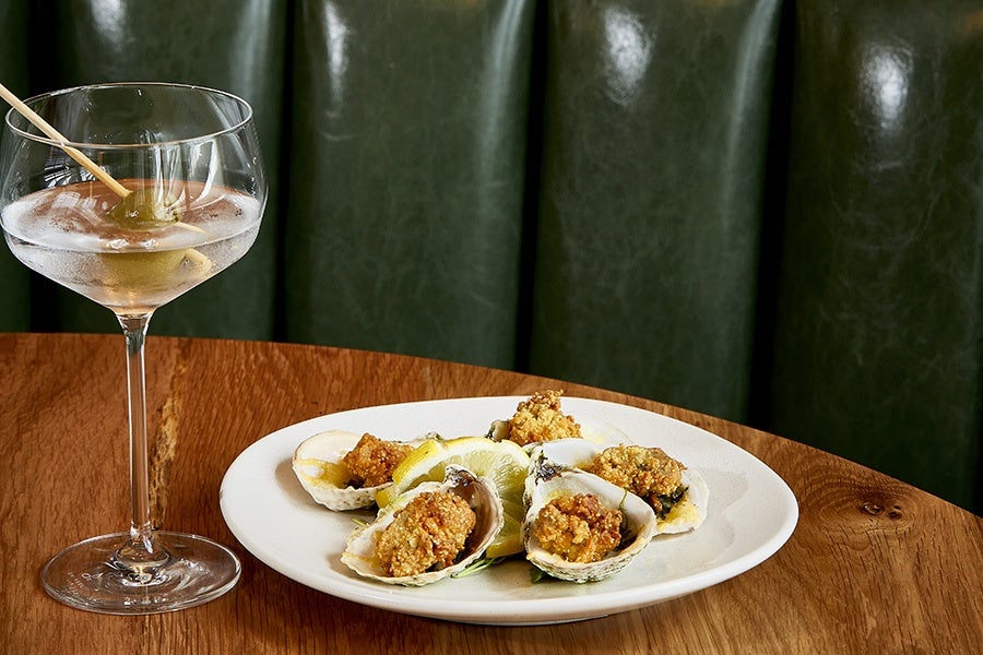 A white plate with six fried oysters garnished with lemon wedges on a wooden table, accompanied by a cocktail glass with a green olive on a skewer. The background features dark green upholstered seating.