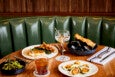 A wooden table set with various dishes and drinks, including a plate of grilled fish with sauce and fries, a bowl of mussels with toasted bread, a plate of fried oysters with lemon wedges, a bowl of grilled asparagus, a glass of iced cocktail with a straw, and a martini glass with olives on a skewer. The background features a green leather booth and a wooden paneled wall. Silverware is placed on a white napkin with black stripes.