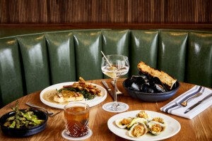A wooden table set with various dishes and drinks, including a plate of grilled fish with sauce and fries, a bowl of mussels with toasted bread, a plate of fried oysters with lemon wedges, a bowl of grilled asparagus, a glass of iced cocktail with a straw, and a martini glass with olives on a skewer. The background features a green leather booth and a wooden paneled wall. Silverware is placed on a white napkin with black stripes.