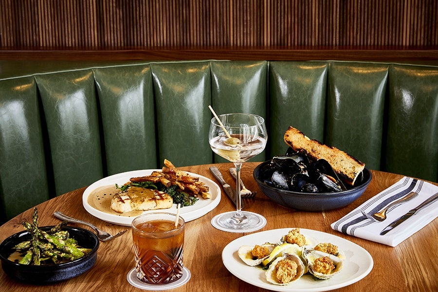 A wooden table set with various dishes and drinks, including a plate of grilled fish with sauce and fries, a bowl of mussels with toasted bread, a plate of fried oysters with lemon wedges, a bowl of grilled asparagus, a glass of iced cocktail with a straw, and a martini glass with olives on a skewer. The background features a green leather booth and a wooden paneled wall. Silverware is placed on a white napkin with black stripes.
