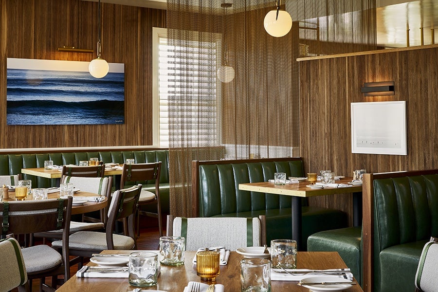 Restaurant interior with green leather booth seating and wooden tables set with glassware, plates, and napkins. The walls are covered in wood paneling, featuring a large photograph of ocean waves and a white framed artwork. Round pendant lights hang from the ceiling, and a sheer brown curtain partially divides the space.