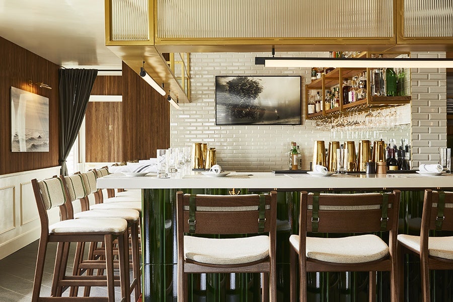 A modern bar area with a white countertop and green glossy base. There are wooden bar stools with light beige cushions arranged along the counter. The back wall features white subway tiles, a mounted TV displaying an abstract image, and wooden shelves holding various bottles and glassware. The ceiling has a gold-framed light fixture, and the left wall is wood-paneled with a framed black-and-white photograph and a dark curtain partially covering a window.