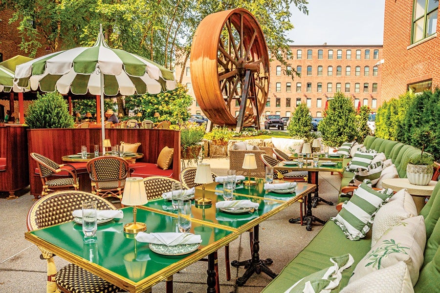 Outdoor dining area with green tables, wicker chairs, and green cushioned bench seating adorned with striped and patterned pillows. A large vintage water wheel is visible in the background, surrounded by greenery and brick buildings. A green and white striped umbrella shades one of the tables.