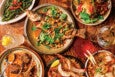 A wooden table set with a variety of dishes and drinks. In the center, a whole cooked fish topped with fresh herbs and cashews on a round plate. Surrounding it are plates with grilled lamb chops in sauce, sautéed greens garnished with sliced red chili, a bowl of rice with shrimp and cashews, and a noodle dish with fresh basil and cherry tomatoes. There are also three drinks: a clear cocktail with olives, a red drink with ice, and a light-colored drink with a pineapple garnish.