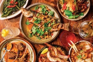 A wooden table set with a variety of dishes and drinks. In the center, a whole cooked fish topped with fresh herbs and cashews on a round plate. Surrounding it are plates with grilled lamb chops in sauce, sautéed greens garnished with sliced red chili, a bowl of rice with shrimp and cashews, and a noodle dish with fresh basil and cherry tomatoes. There are also three drinks: a clear cocktail with olives, a red drink with ice, and a light-colored drink with a pineapple garnish.