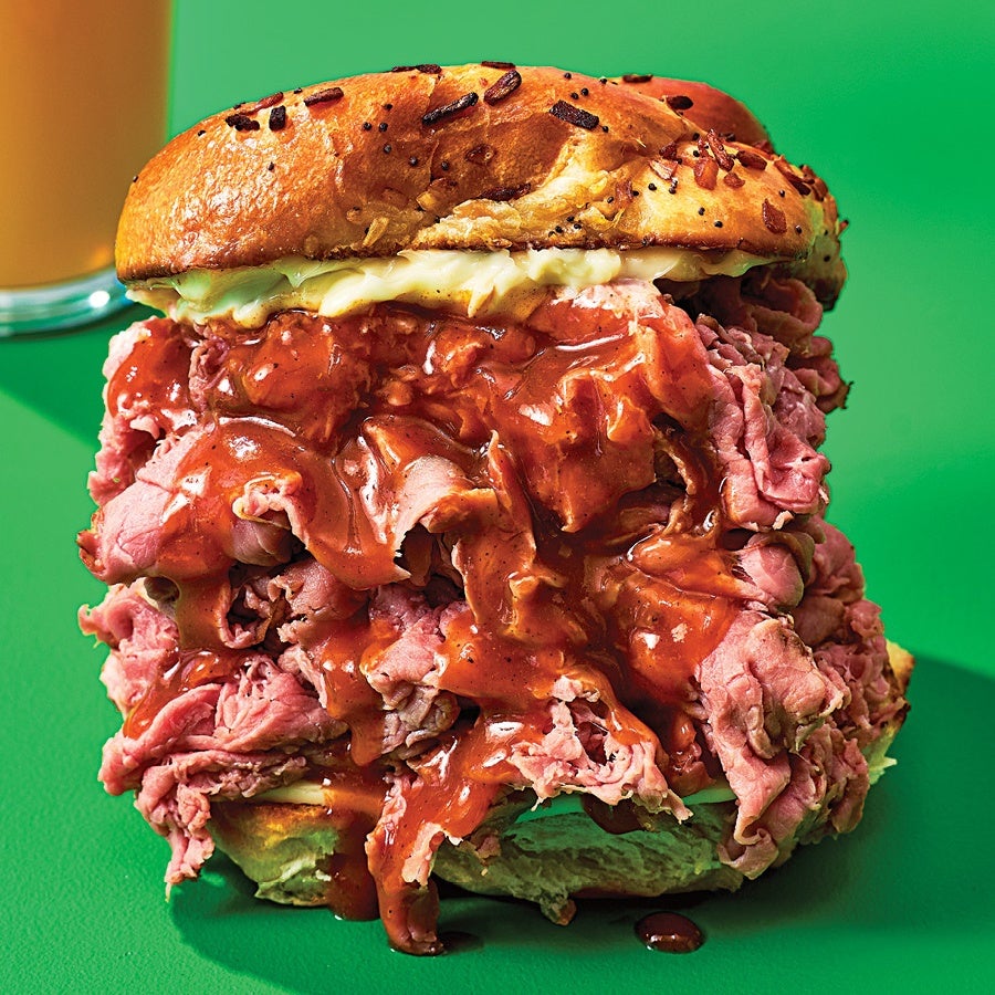 A sandwich with a toasted onion bun filled with a generous amount of thinly sliced roast beef, topped with a rich, reddish-brown gravy sauce. The sandwich is placed on a green surface.