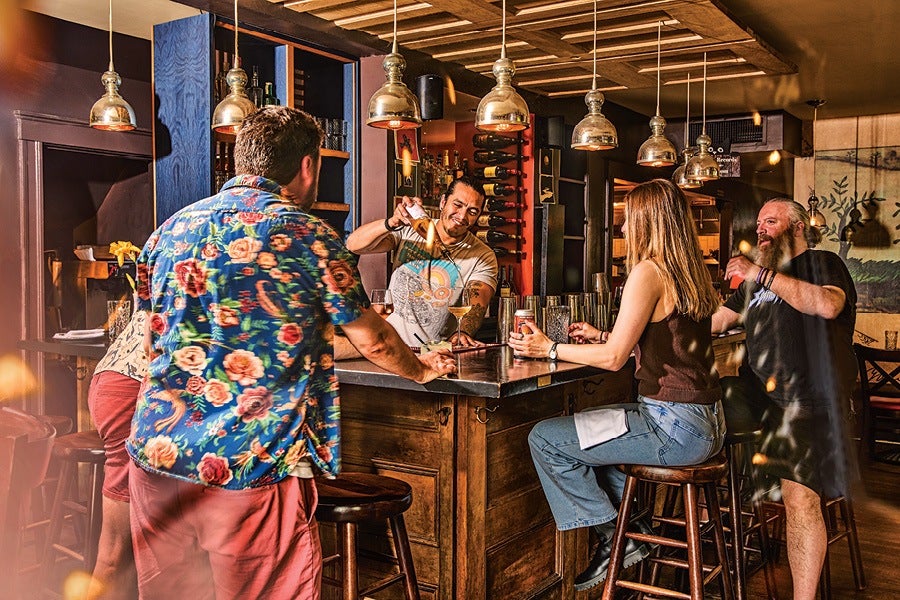 A lively bar scene with a bartender pouring a flaming drink behind a wooden bar counter. Four patrons are seated on stools around the bar, engaged and watching the bartender. The bar has hanging pendant lights and shelves stocked with bottles in the background. The atmosphere is warm and inviting.
