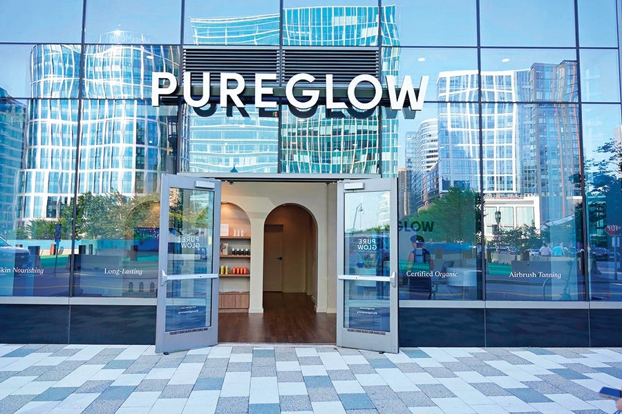 Glass storefront of a business named "PUREGLOW" with large white letters above the entrance. The double glass doors are open, revealing a clean interior with wooden floors, shelves with products, and an arched doorway inside. The glass facade reflects modern high-rise buildings and trees across the street. Text on the windows includes phrases like "Skin Nourishing," "Long-Lasting," "Certified Organic," and "Airbrush Tanning." The sidewalk in front is paved with a pattern of light and dark rectangular tiles.