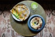 A round table with a plate of flatbreads topped with squash blossoms, a blue bowl of creamy hummus garnished with olive oil, crumbled cheese, and spices, and a glass of green juice. The table is set on a patterned rug.