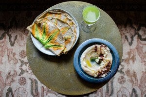 A round table with a plate of flatbreads topped with squash blossoms, a blue bowl of creamy hummus garnished with olive oil, crumbled cheese, and spices, and a glass of green juice. The table is set on a patterned rug.