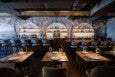 Modern restaurant interior with wooden tables set with plates, wine glasses, and cutlery. The bar area features high stools, illuminated circular shelves with bottles, glasses, and plates, and a decorative backdrop with tree and bird motifs. Warm lighting creates a cozy atmosphere.