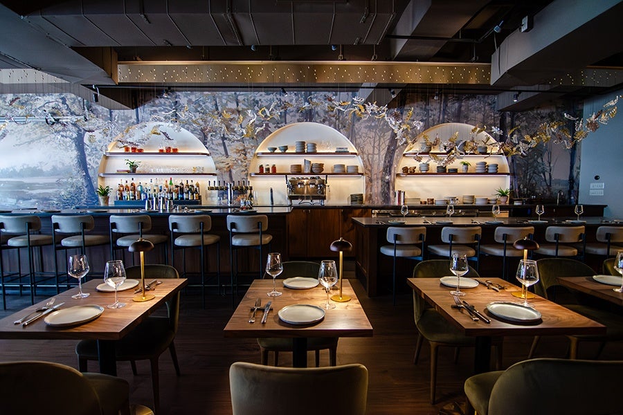 Modernes Restaurantinterieur mit Holztischen, gedeckt mit Tellern, Weingläsern und Besteck. Der Barbereich verfügt über hohe Hocker, beleuchtete runde Regale mit Flaschen, Gläsern und Tellern sowie eine dekorative Kulisse mit Baum- und Vogelmotiven. Warmes Licht schafft eine gemütliche Atmosphäre.
