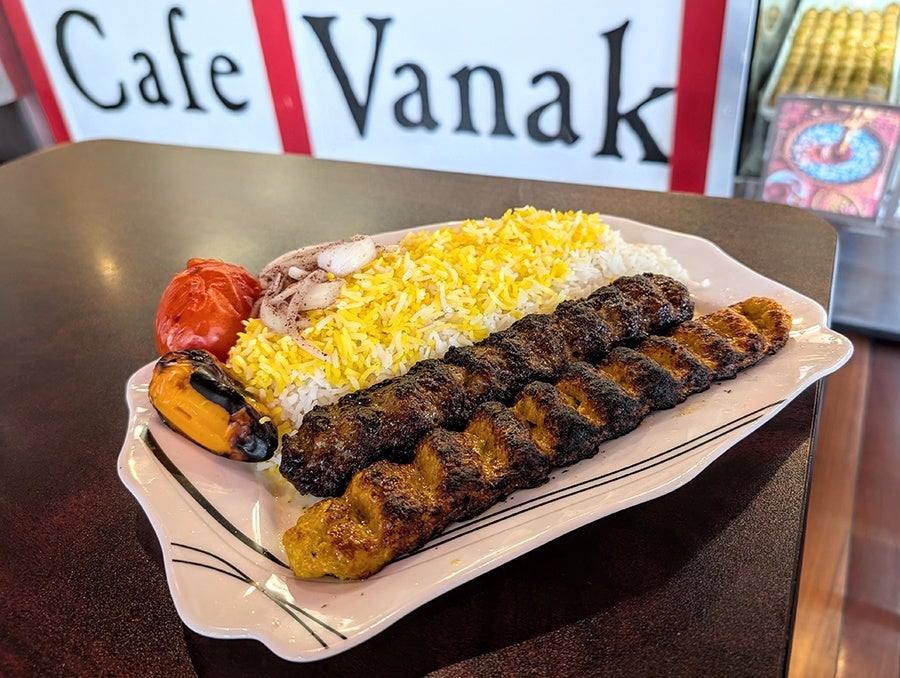 Kebabs and yellow-and-white rice, with roasted peppers and tomatoes, sit on a white plate in front of a counter with large lettering reading Cafe Vanak.