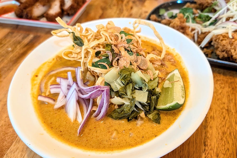 A bowl of yellow soup is topped with crispy noodles, a lime wedge, pickled mustard greens, and slivers of raw red onions.