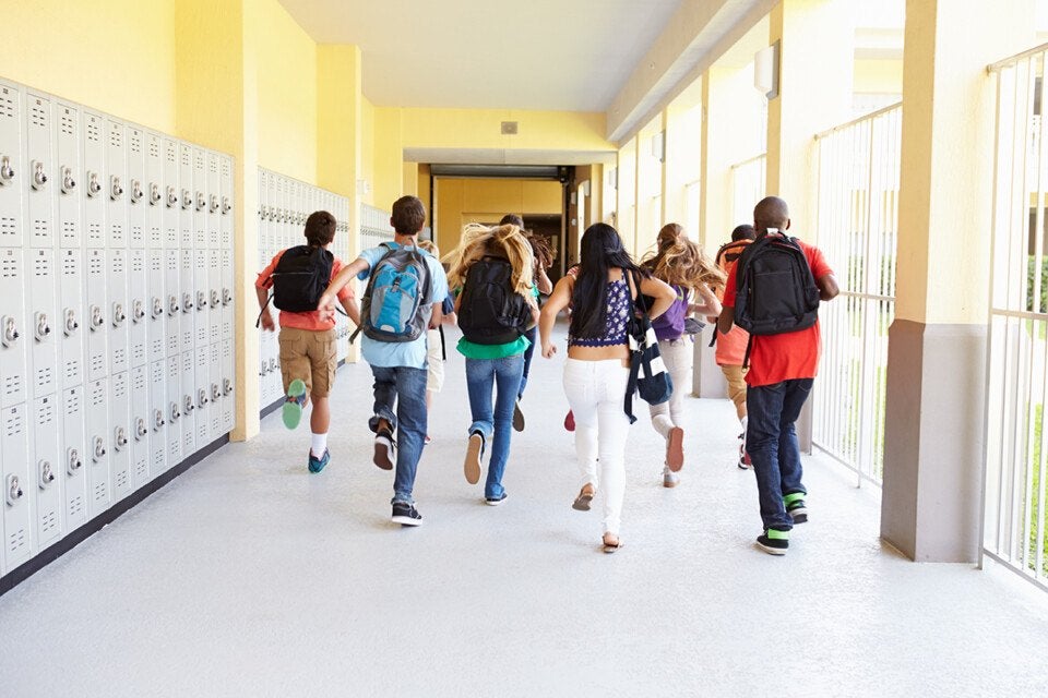 A group of students with backpacks running down a school hallway lined with lockers on the left and large windows on the right. The walls are painted yellow and the floor is light-colored. The students are seen from behind.