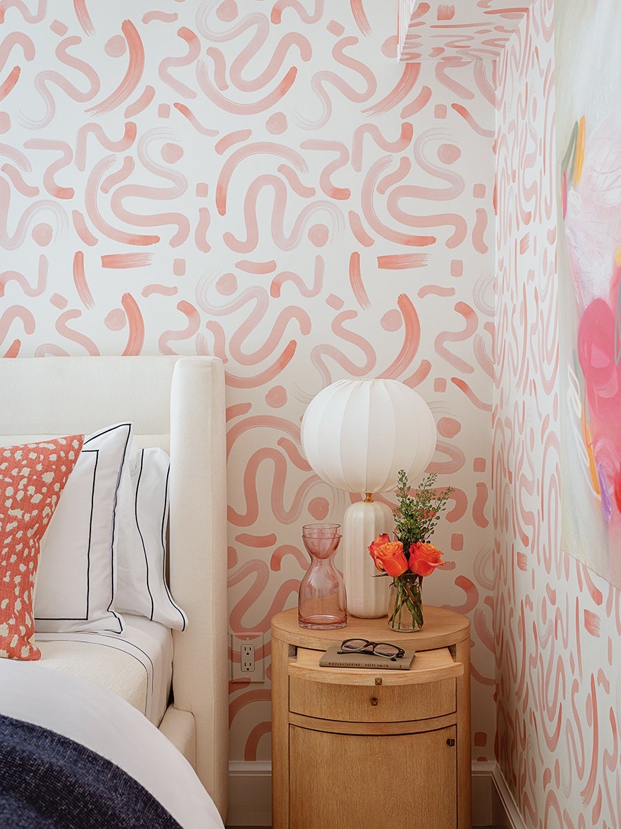 A bedroom corner featuring a light beige upholstered bed with white pillows outlined in black and a coral-patterned accent pillow. Next to the bed is a round wooden nightstand holding a white, round, pleated table lamp, a pink glass vase, a small bouquet of orange roses with greenery in a clear glass jar, and a pair of black-rimmed glasses resting on a book. The walls are decorated with a white and coral abstract squiggle pattern.