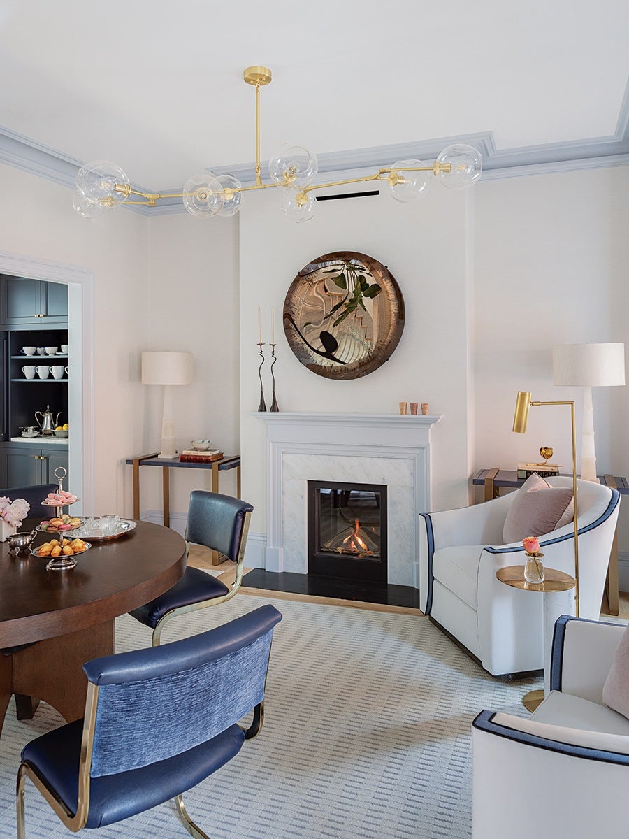 Modern living room with a white marble fireplace, a round wooden dining table with blue upholstered chairs, and two white armchairs with navy trim and pink cushions. The room features a light gray patterned carpet, a gold and glass chandelier, a round decorative mirror above the fireplace, and gold-accented floor and table lamps. A small side table with a pink flower in a vase is next to one armchair, and a corner table with a lamp and books is visible. The adjacent kitchen area has dark cabinetry with white cups and a silver teapot on display.