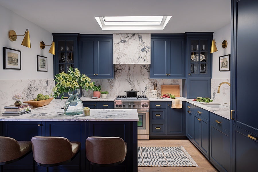 Kitchen with navy blue cabinets and drawers, white marble countertops and backsplash, a stainless steel stove with a black pot on it, and a skylight above. The island has a clear glass vase with green and white flowers, three brown leather bar stools, and a few books. There are gold wall sconces and matching gold cabinet handles and faucet. A patterned rug is on the wooden floor in front of the stove.