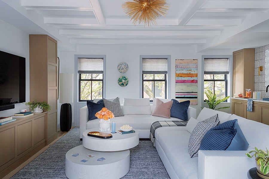 Modern living room with white sectional sofas adorned with blue, gray, and pink throw pillows. Two round white nested coffee tables hold a bouquet of orange roses, candles, and a bowl of popcorn. The room features three windows with light gray blinds, light wood cabinetry, a large flat-screen TV, and a gray area rug. A gold starburst chandelier hangs from the coffered ceiling, and colorful wall art and plants add decorative touches.