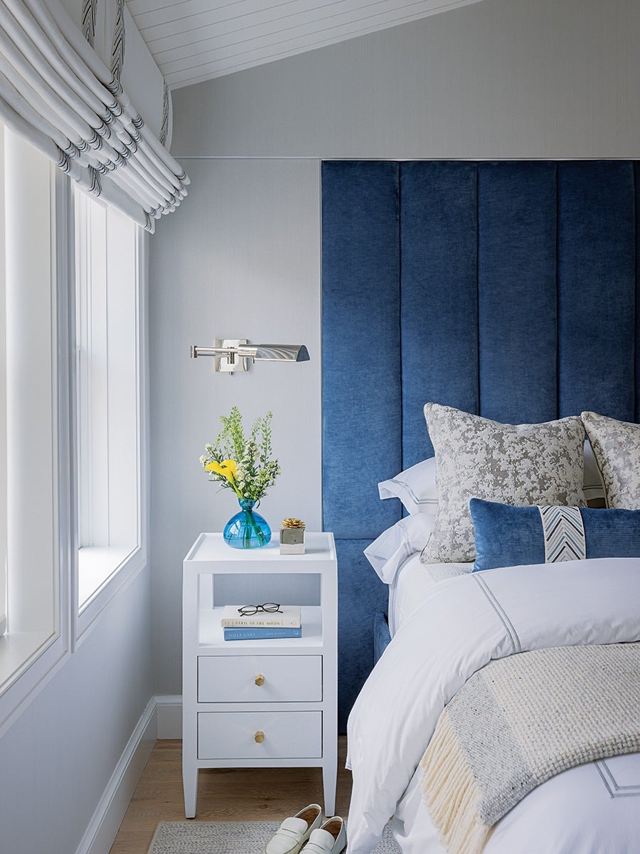 A modern bedroom corner featuring a white nightstand with two drawers and an open shelf holding two books and a pair of glasses. On top of the nightstand is a blue glass vase with yellow and green flowers and a small decorative item. The bed has a tall, upholstered blue headboard and is dressed in white bedding with a textured beige throw blanket and several pillows in shades of blue and gray. A silver wall-mounted reading light is above the nightstand. The room has light gray walls, a light wood floor, and large windows with white Roman shades. White slippers are placed on a light gray rug next to the nightstand.
