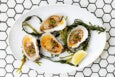 Four dressed oysters topped with a golden sauce and garnished with sliced green herbs, served on a white oval plate with green seaweed and a lemon wedge, placed on a white hexagonal tile surface.
