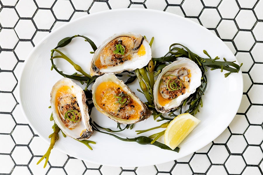 Four dressed oysters topped with a golden sauce and garnished with sliced green herbs, served on a white oval plate with green seaweed and a lemon wedge, placed on a white hexagonal tile surface.