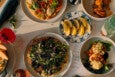 Overhead view of restaurant dishes on a white marble table with dusk-like lighting. There's a salad, a flatbread, a plate of lemon wedges, a pink cocktail, and more.
