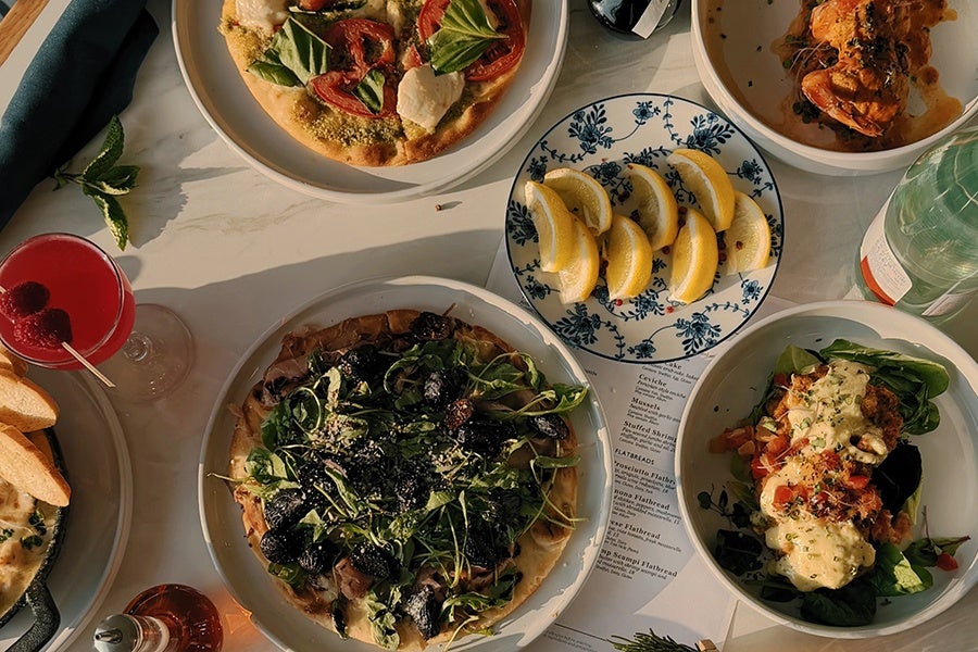 Overhead view of restaurant dishes on a white marble table with dusk-like lighting. There's a salad, a flatbread, a plate of lemon wedges, a pink cocktail, and more.
