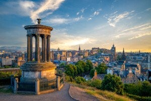 A stone monument with tall columns and a circular roof stands on a hill overlooking a cityscape at sunset. The city features historic buildings, trees, and a clock tower, with a partly cloudy sky glowing with warm light.