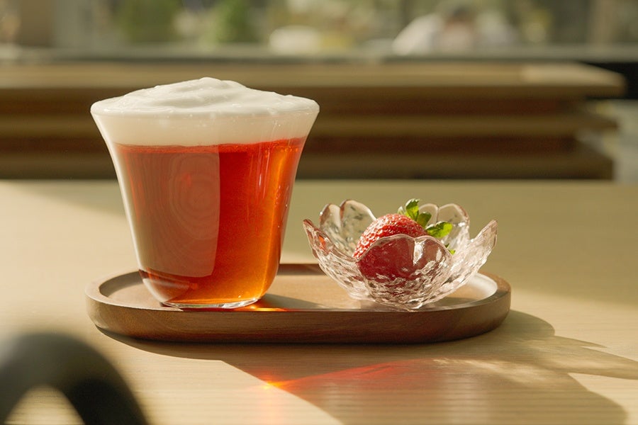 A glass of frothy tea with a rich amber color sits on a wooden tray next to a small, clear, flower-shaped glass bowl holding a single ripe strawberry with green leaves. The scene is softly lit with natural light.