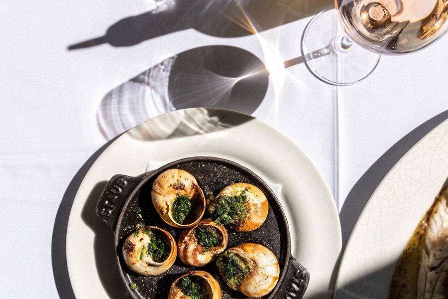 A black cast iron dish containing six cooked escargots topped with green herb butter, placed on a white plate. To the upper right, there is a glass of rosé wine casting a shadow on the white tablecloth. Part of another white plate with food is visible on the right edge.