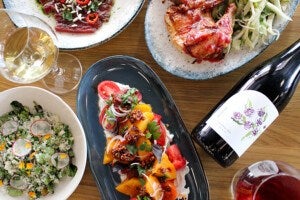 A wooden table set with various dishes and drinks. There is a plate of grilled chicken with a side of sliced fennel, a bowl of green salad topped with radish slices and edible flowers, a dark oval plate with a colorful tomato salad garnished with herbs and onions, and a plate of thinly sliced raw meat topped with chili and herbs. A glass of white wine and a glass of red wine are also visible, along with a bottle of red wine with a white label featuring a floral design.