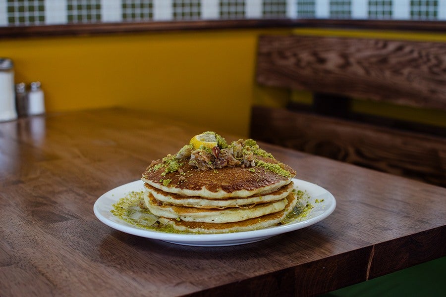 A stack of three golden-brown pancakes on a white plate, topped with chopped nuts, a lemon slice, and green crumbled garnish, placed on a wooden table with a yellow wall and wooden bench in the background.