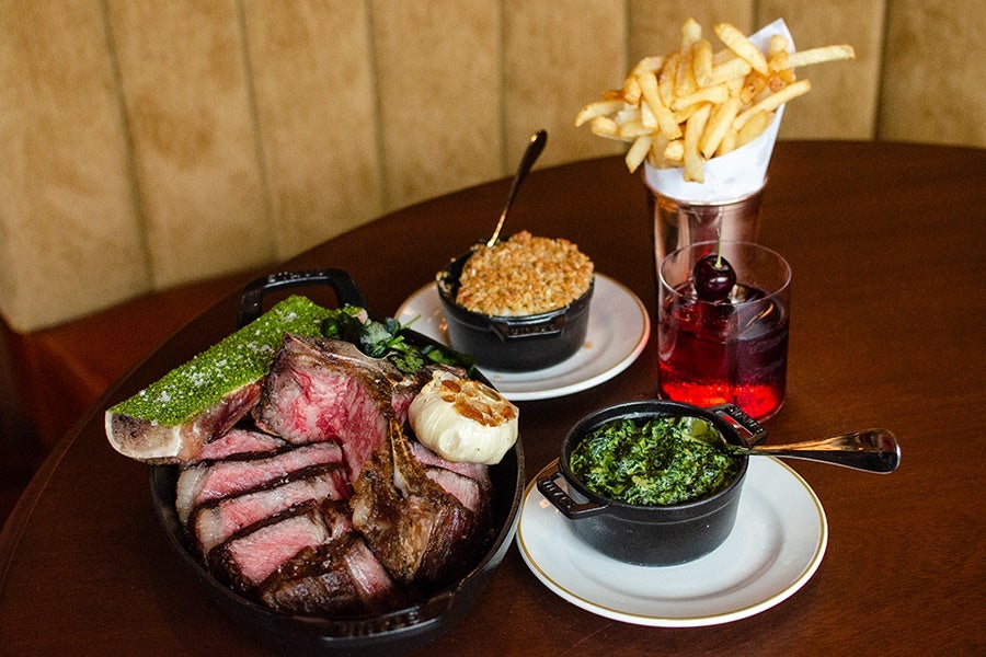 A meal featuring a cast iron skillet with sliced medium-rare steak, a bone with green herb butter, and a roasted garlic bulb. On the side, there is a small black pot of creamed spinach on a white plate, a small black pot of baked macaroni and cheese with a spoon, a glass of red cocktail garnished with a cherry, and a metal cup filled with French fries wrapped in white paper. All items are placed on a dark wooden table.