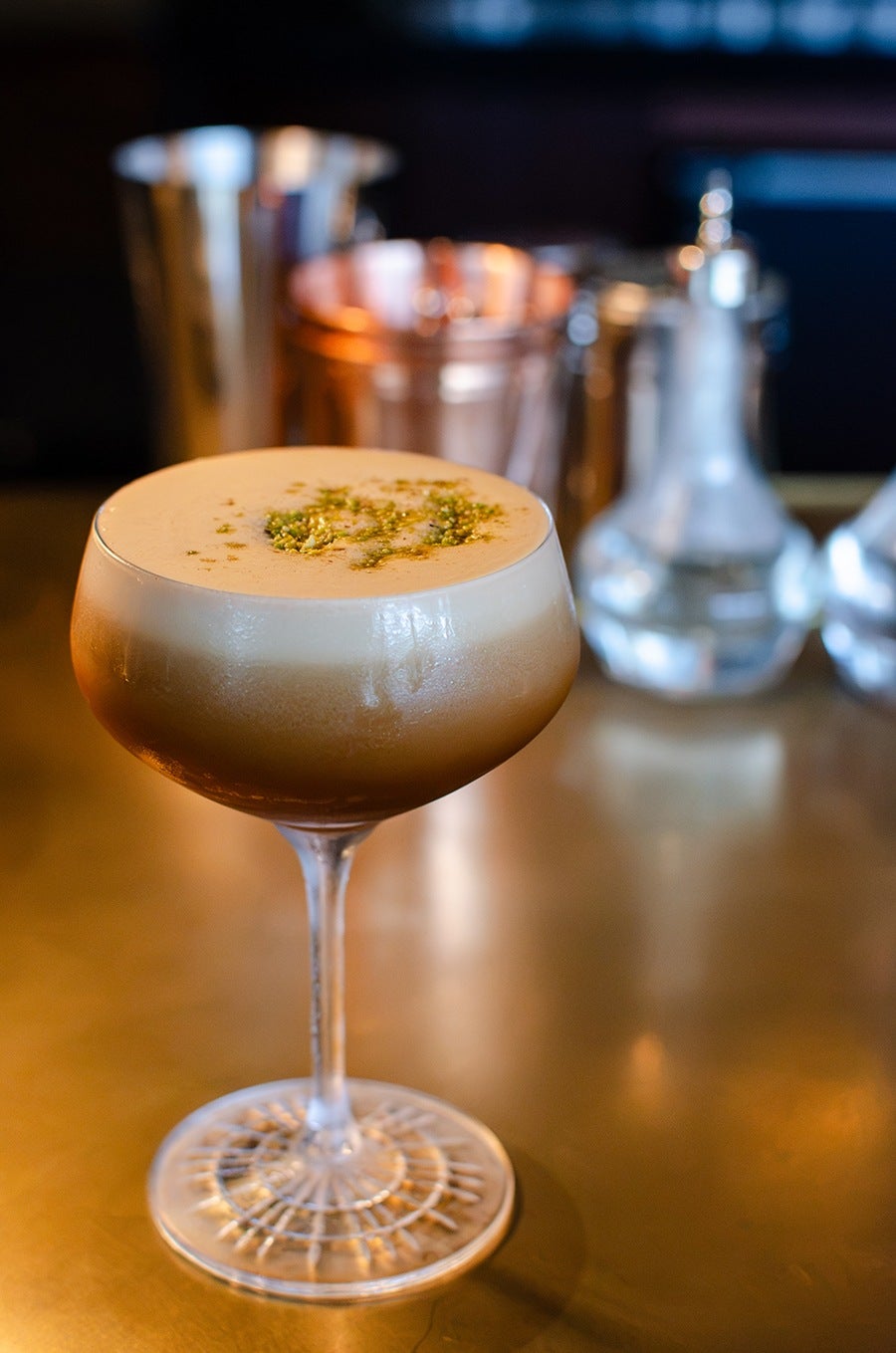 A cocktail in a coupe glass with a frothy, creamy top layer, garnished with a sprinkle of green herbs or spices. The glass is placed on a reflective golden surface, with blurred metallic bar tools and glass bottles in the background.