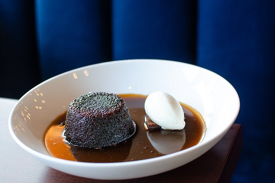 A sticky toffee pudding served in a white bowl with a pool of caramel sauce, accompanied by a scoop of vanilla ice cream on the side.